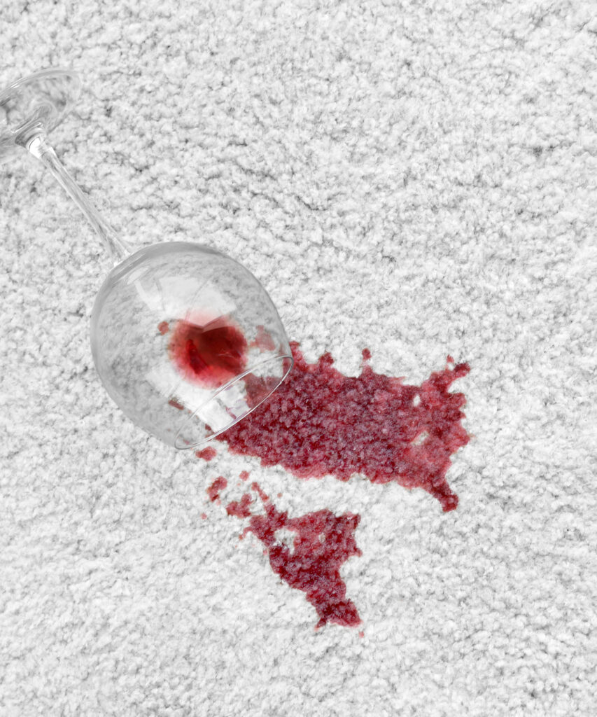 Glass of red wine spilled on fluffy white carpet, leaving behind a large red stain. 