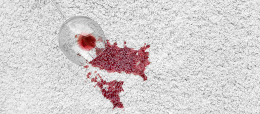 Glass of red wine spilled on fluffy white carpet, leaving behind a large red stain. 